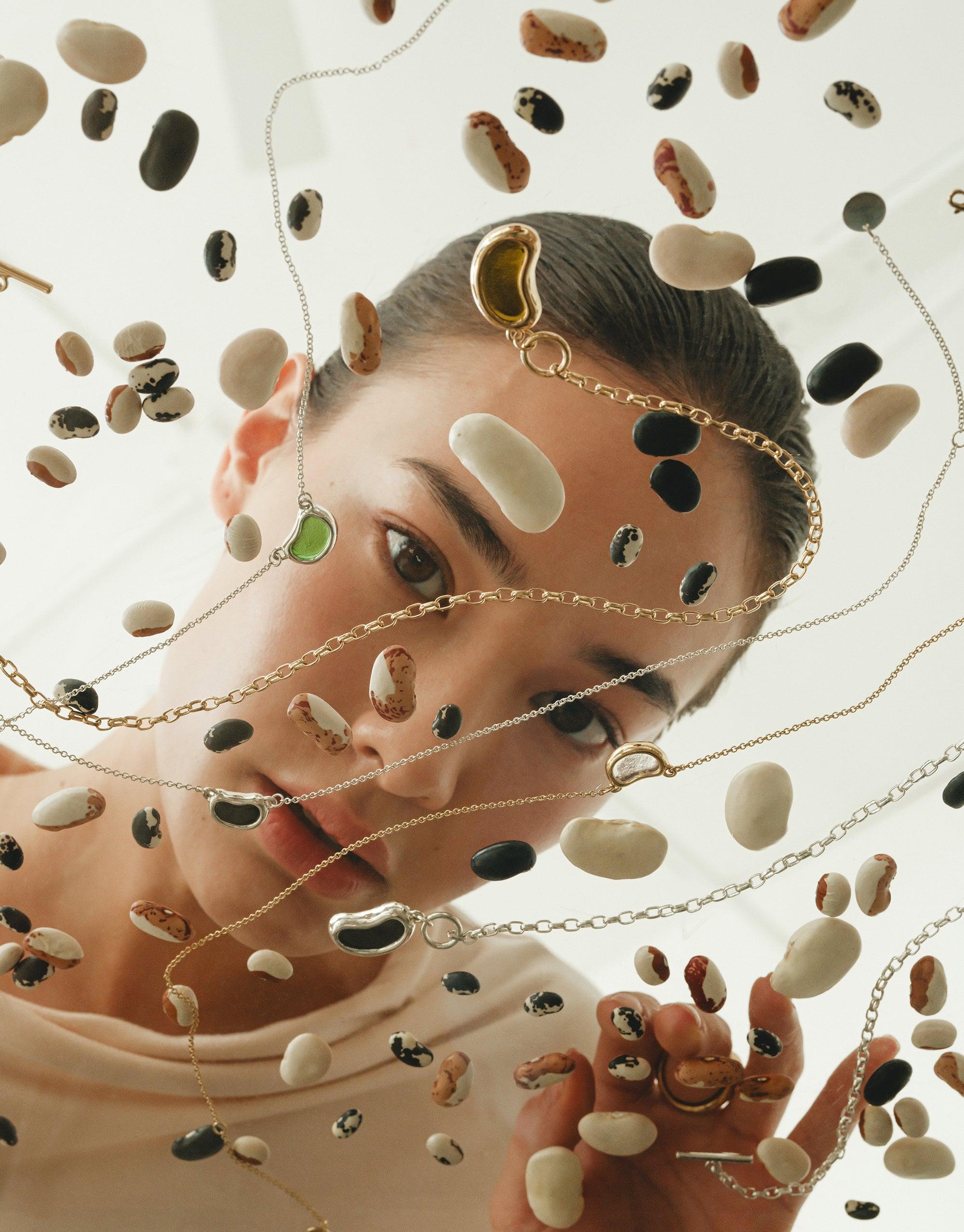 A woman peers through a glass adorned with multiple necklaces with spread beans, showcasing eco-conscious jewelry, handmade from repurposed glass, reflecting CLED's commitment to sustainability.