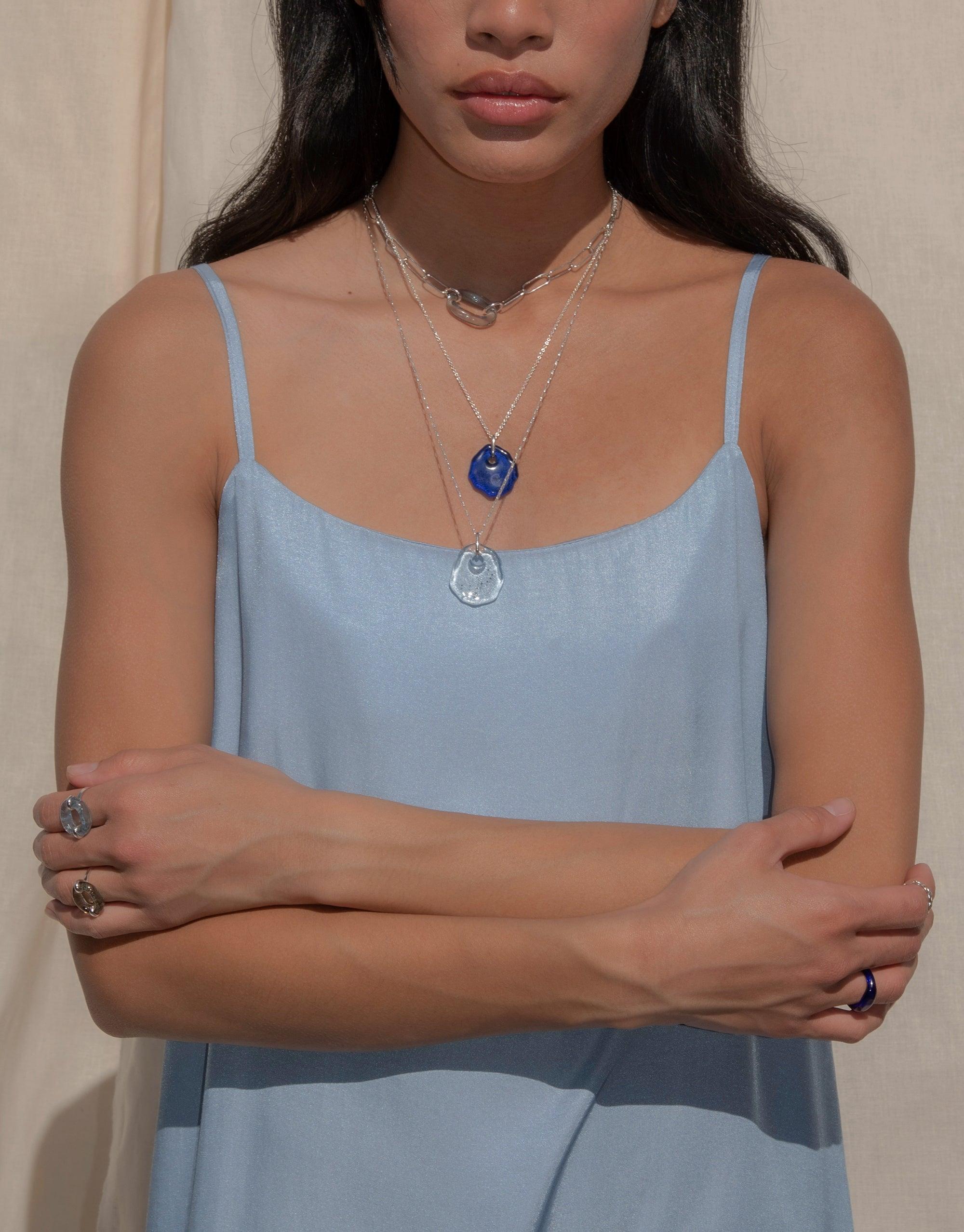 The Day Loop Necklace features a sleek oval chain with a hand-formed Eco Gem centerpiece, worn by a woman in a blue dress, showcasing its minimalist elegance and eco-conscious design.