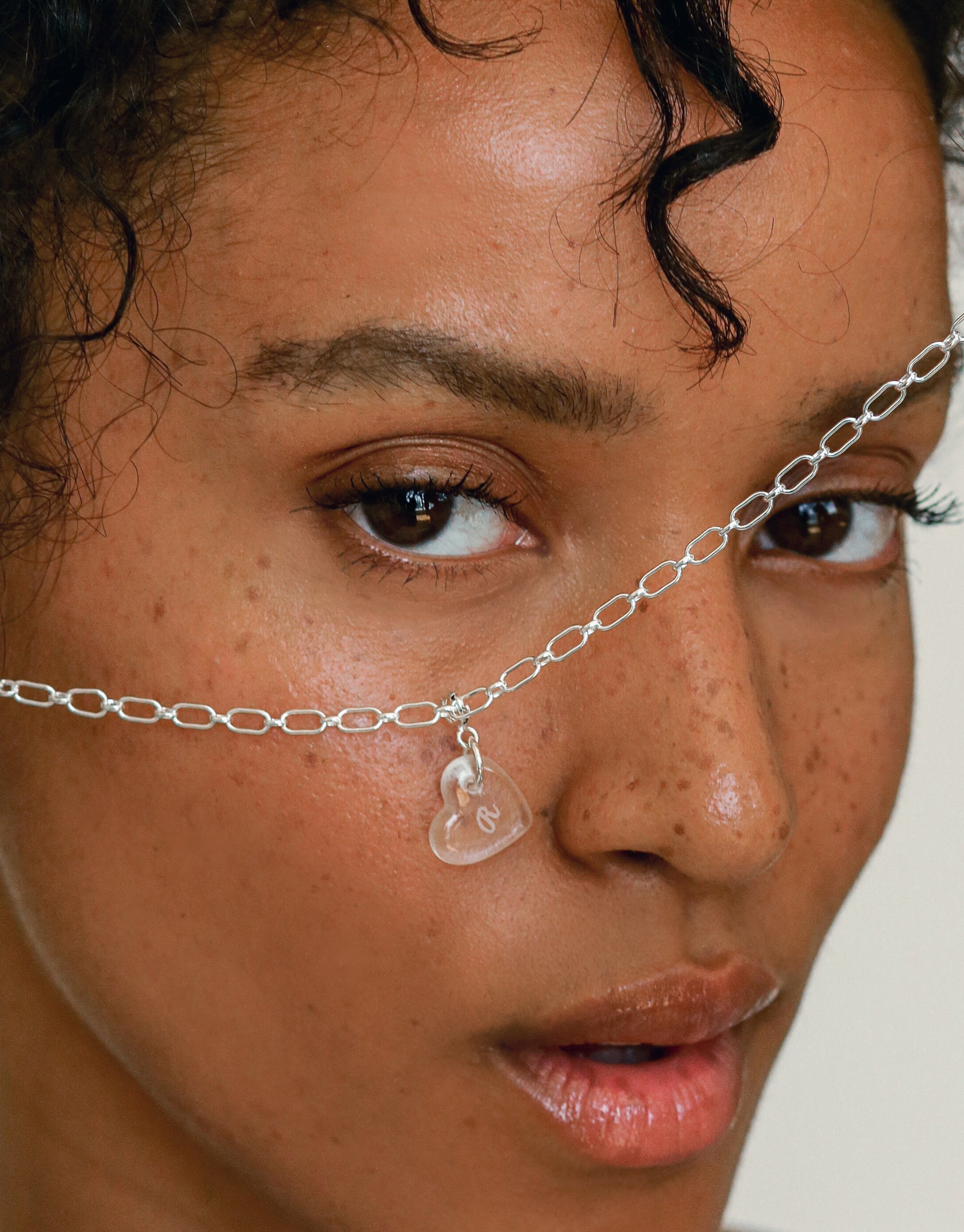 Close-up of a delicate silver chain necklace featuring a transparent heart-shaped charm. The necklace rests gently across the bridge of the nose, highlighting the Heart Icon Charm with &quot;R&quot; engraving. Handcrafted jewelry by CLED.